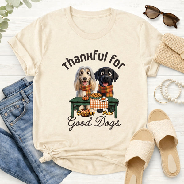 A sand-colored graphic t-shirt styled in a flat lay with blue jeans. The t-shirt features a graphic of custom dogs sitting at a table with the text 'Thankful for Good Dogs' and custom with dog's names
