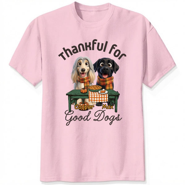 A pink short-sleeve crewneck t-shirt featuring a graphic print of custom dogs sitting at a table with autumn food, with the text 'Thankful for Good Dogs' and custom dog's names written around the illustration.