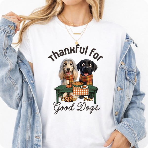 A woman wearing a white graphic t-shirt with a cartoon dog design and text. The graphic features custom dogs sitting at a table with the text 'Thankful for Good Dogs'.