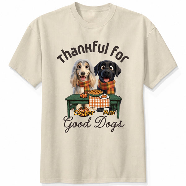 A sand short-sleeve crewneck t-shirt featuring a graphic print of custom dogs sitting at a table with autumn food, with the text 'Thankful for Good Dogs' and custom dog's names written around the illustration.